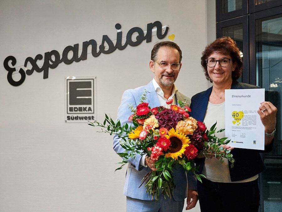 Mann mit Blumenstrau8 steht neben einer Frau mit Urkunde