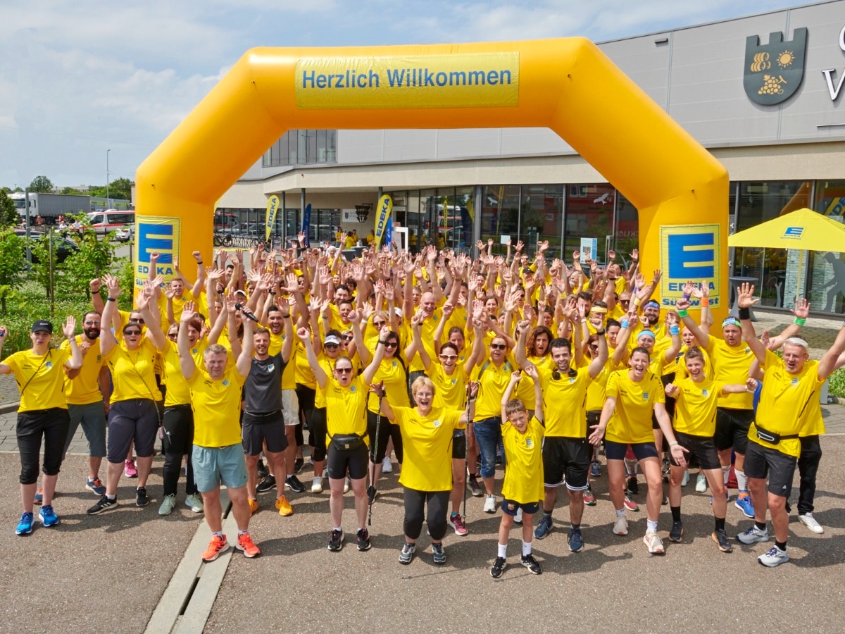 Rund 200 Mitarbeiterinnen und Mitarbeiter tragen ein gelbes Laufschirt mit einem blauen EDEKA Logo, halten die Arme in die Luft  und jubeln gemeinsam