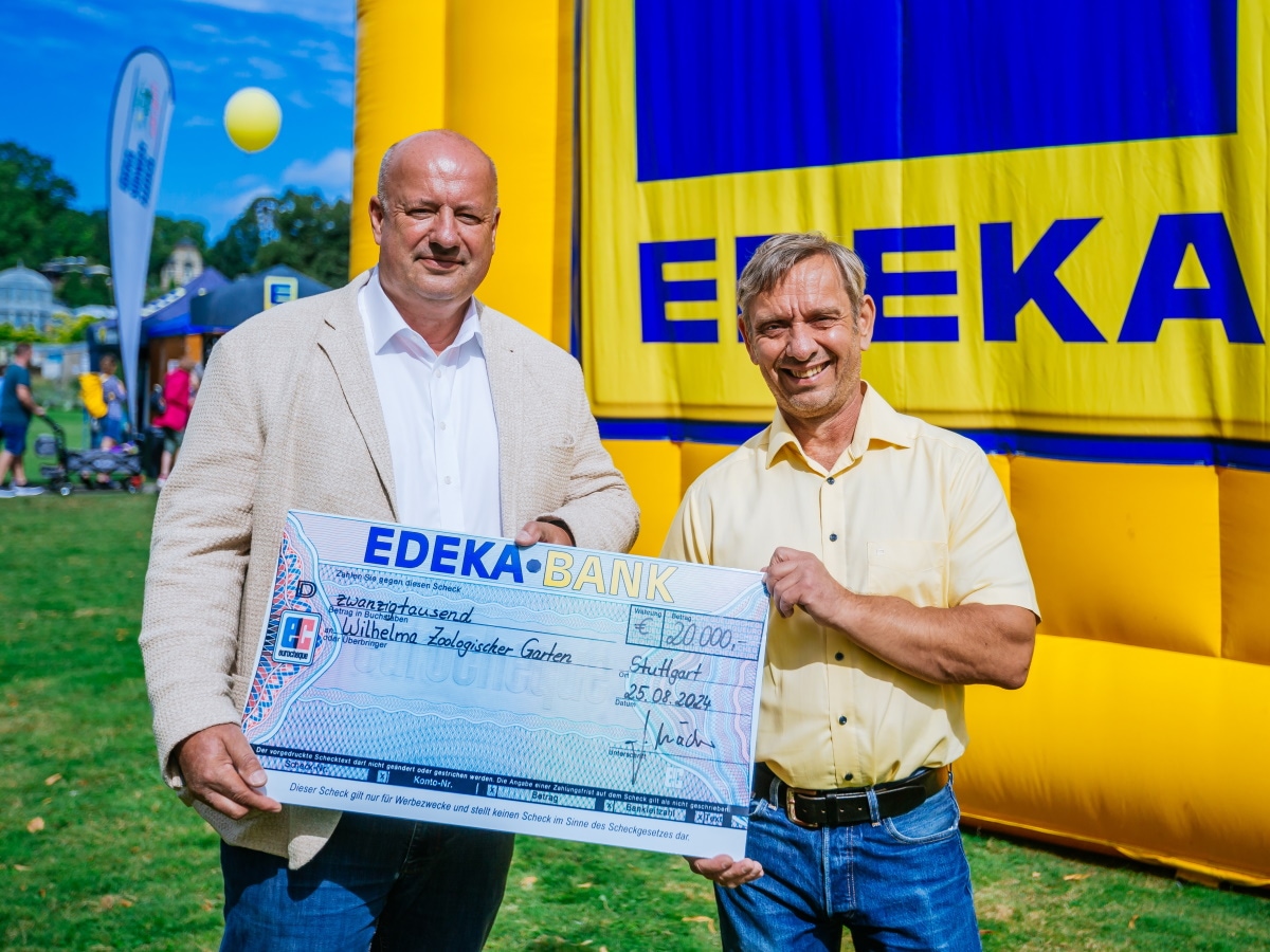 2 Männer stehen in der Sonne und halten gemeinsam einen Scheck mit 20.000 € für die Wilhelma, dahinter eine EDEKA-Hüpfburg