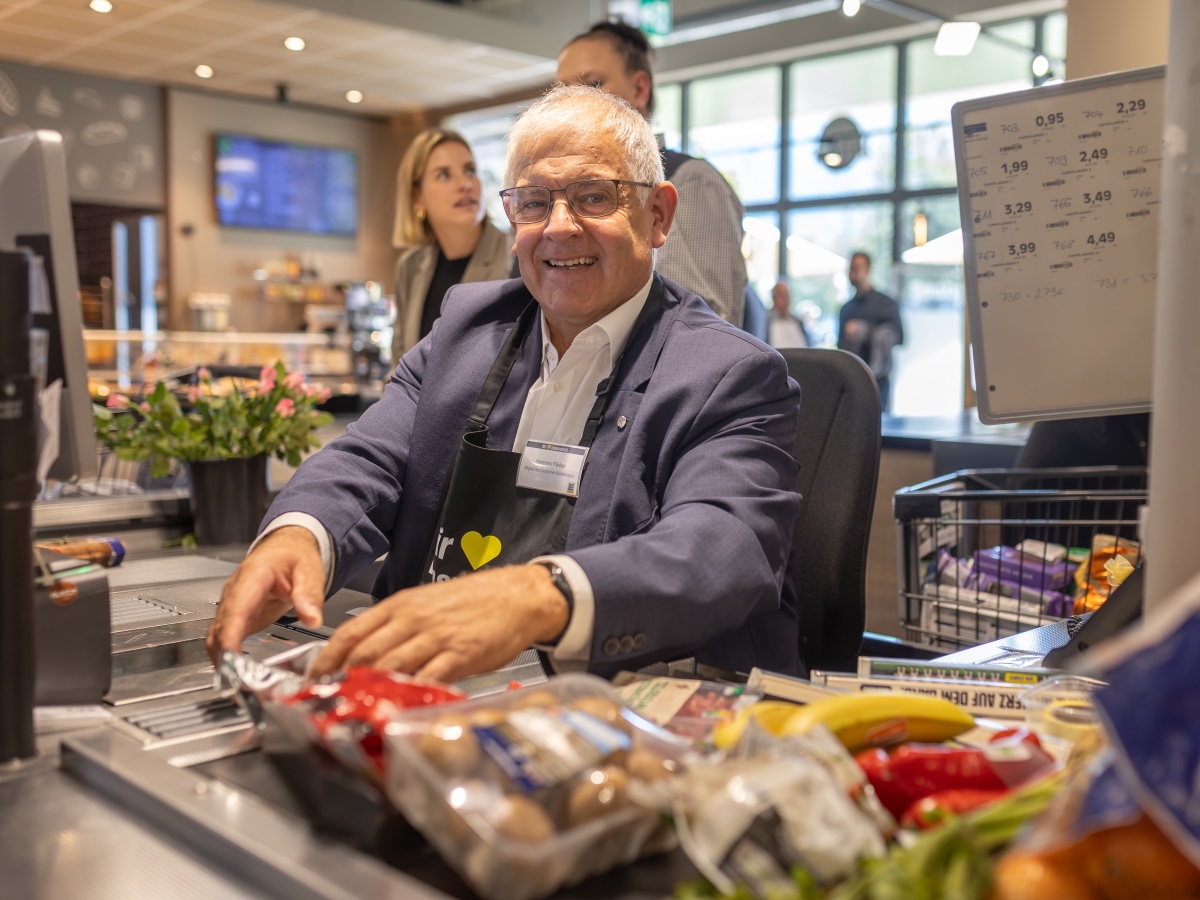 Ein Mann mit Jackett und schwarzer Schürze sitzt an der Supermarktkasse, lächelt und scannt den Einkauf vom Band
