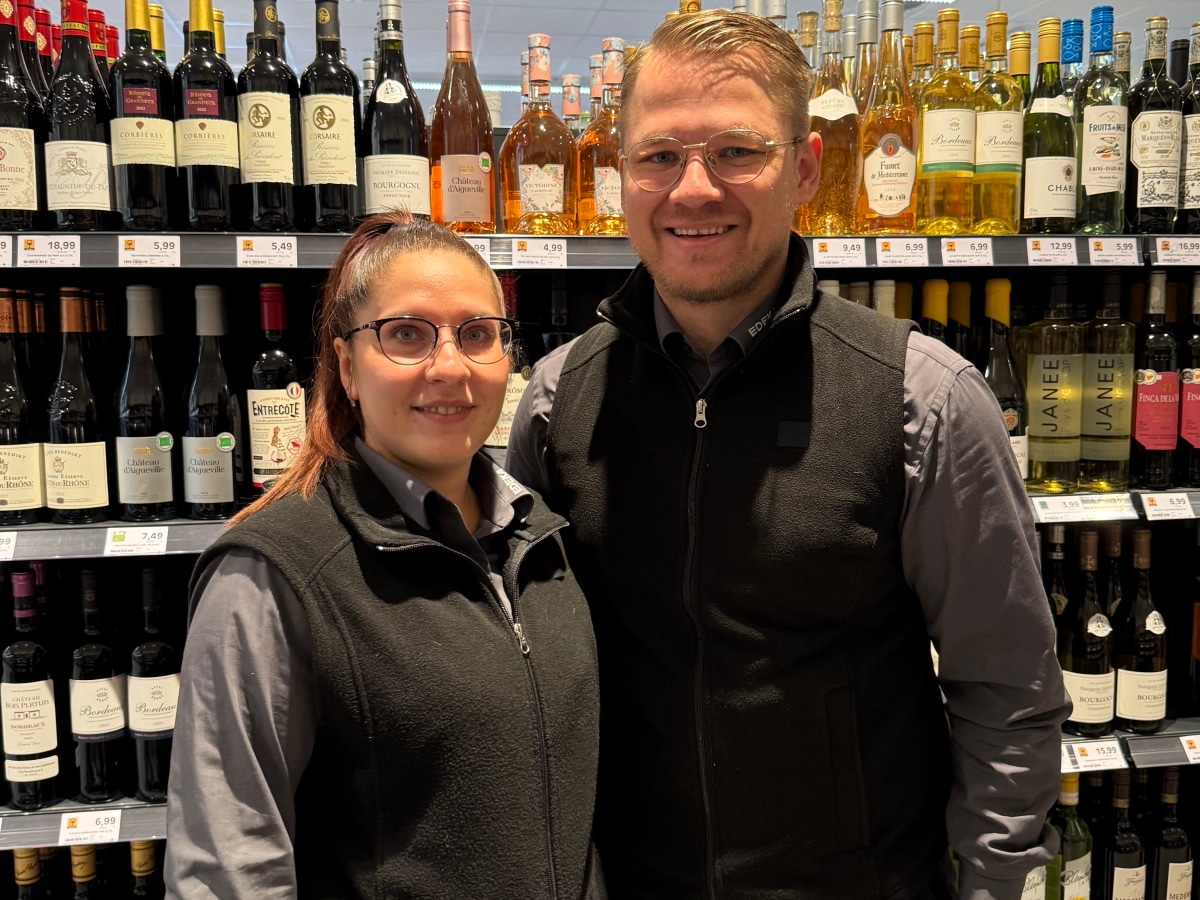 Ein Pärchen mit Arbeitskleidung (graues Hemd und graue Weste) steht im Markt vor einem Weinregal und lächeln