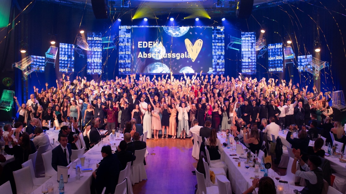 EDEKA Abschlussgala: rund 200 festliche Gekleidete stehen jubelnd zusammen in einem großen Saal mit langen weißen Tischen.