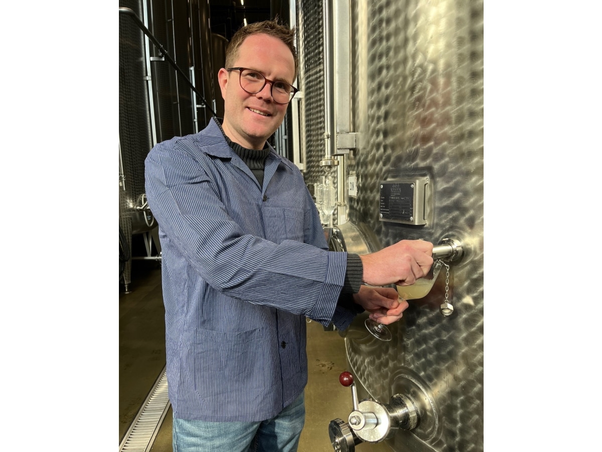 Ein Mann mit Brille und einem blauen Winzerhemd steht vor einem riesigen Stahltank, aus dem er eine Probe in ein Glas abfüllt