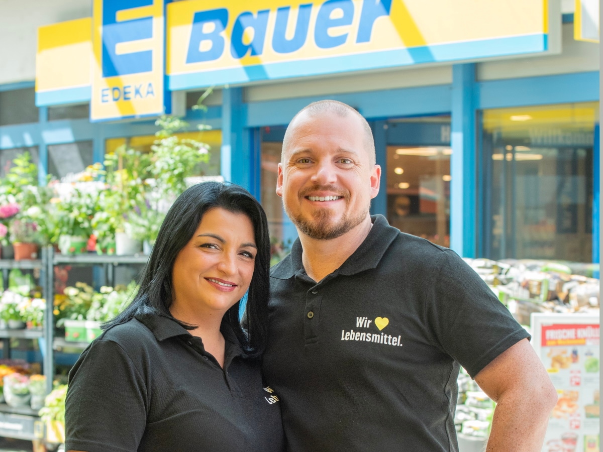 Lächelndes Pärchen mit gleichen T-Shirts steht bei Sonne vorm Eingang eines Marktes. Darüber die Aufschrift EDEKA Bauer