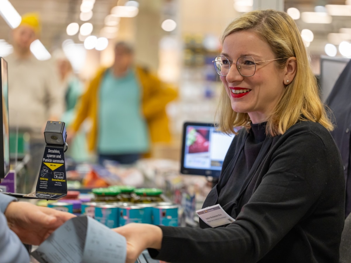 Eine Frau in schwarzer Schürze sitzt an der Supermarktkasse, lächelt und gibt einem Kunden den Kassenzettel.