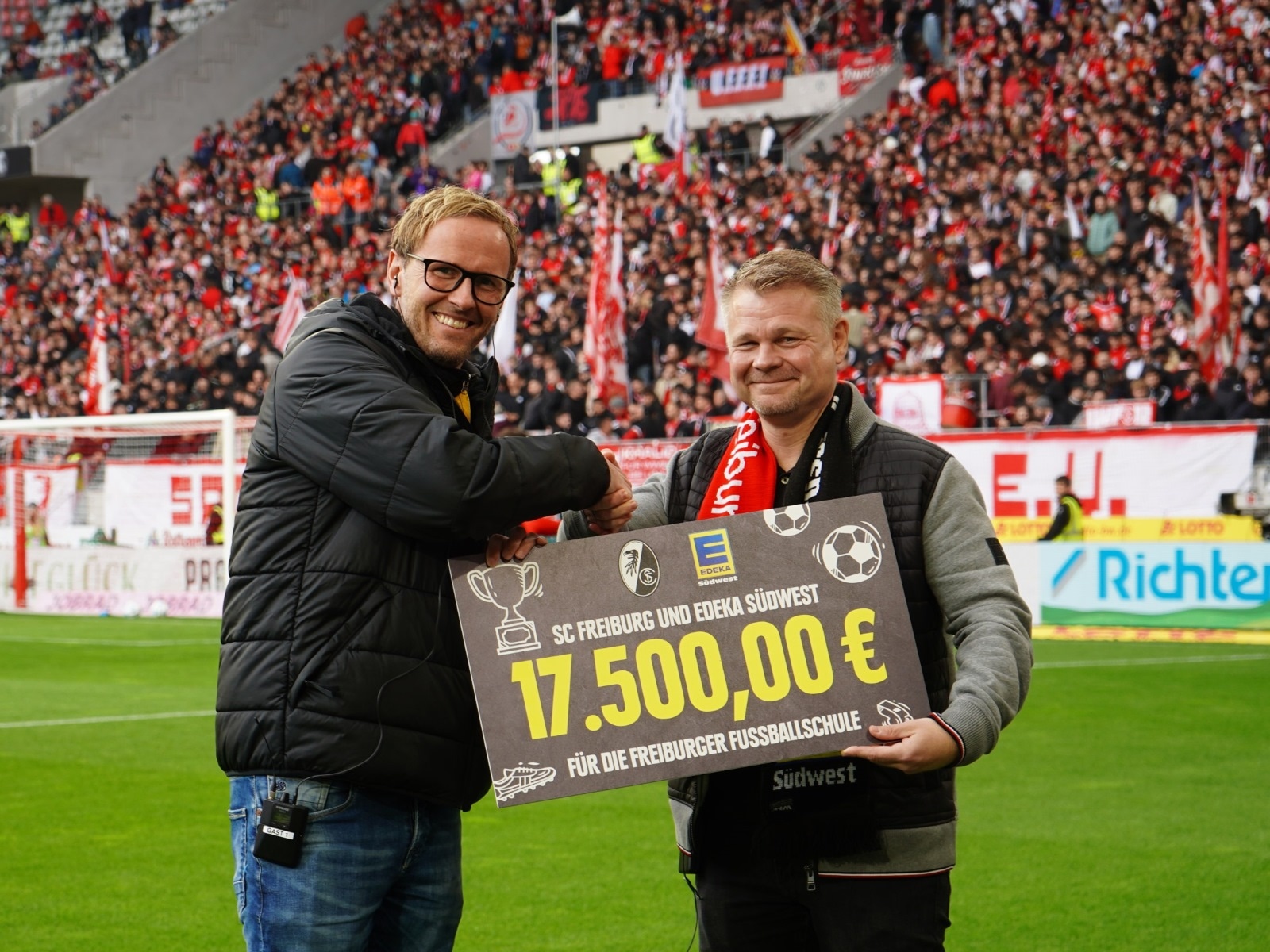 2 Männer mit Schild (17.500 € für die Fußballschule) stehen auf dem Rasen des mit Zuschauern gefüllten SC Freiburg Stadion 