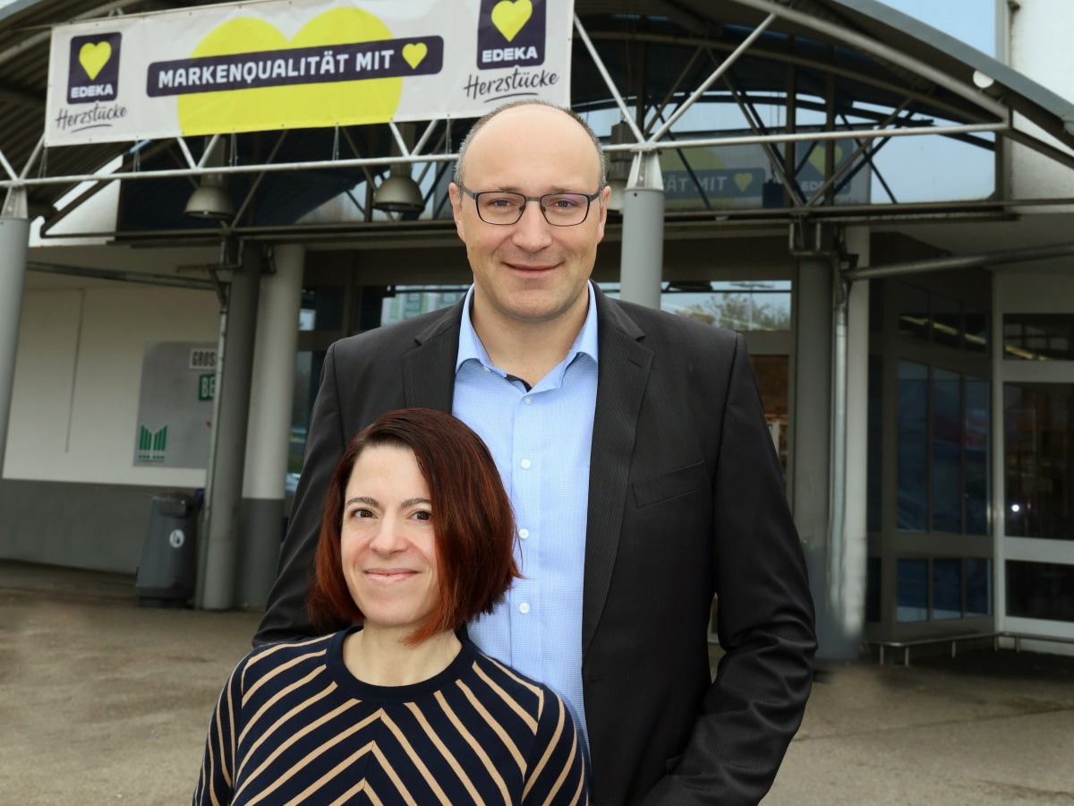 Ein großer Mann und eine kleine Frau stehen zusammen vor dem Eingang eines Marktes, darüber ein EDEKA-Herzstücke-Banner