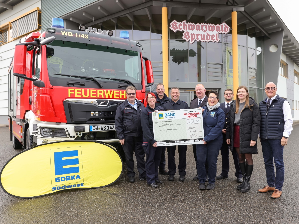 Ein Feuerwehrauto und 9 lachende Personen mit einem großen Scheck stehen vor dem Eingang der Firma Schwarzwald-Sprudel.