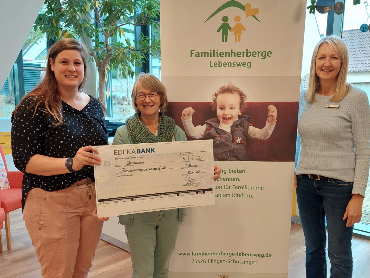 3 strahlende Frauen stehen neben einem Banner mit der Aufschrift Familienherberge Lebensweg und halten einen großen Scheck.