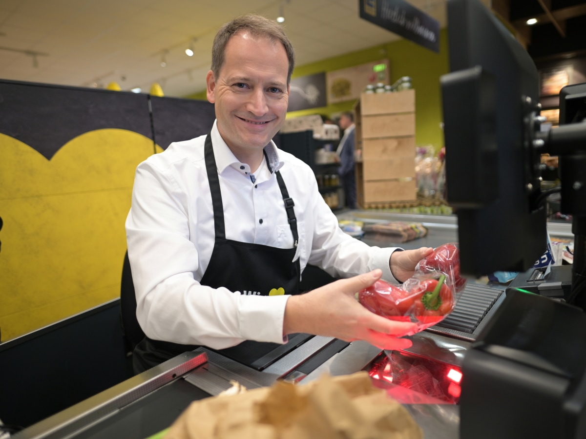 Ein Mann mit weißem Hemd und schwarzer Schürze sitzt an der Supermarktkasse, lächelt und zieht rote Paprika über den Scanner.