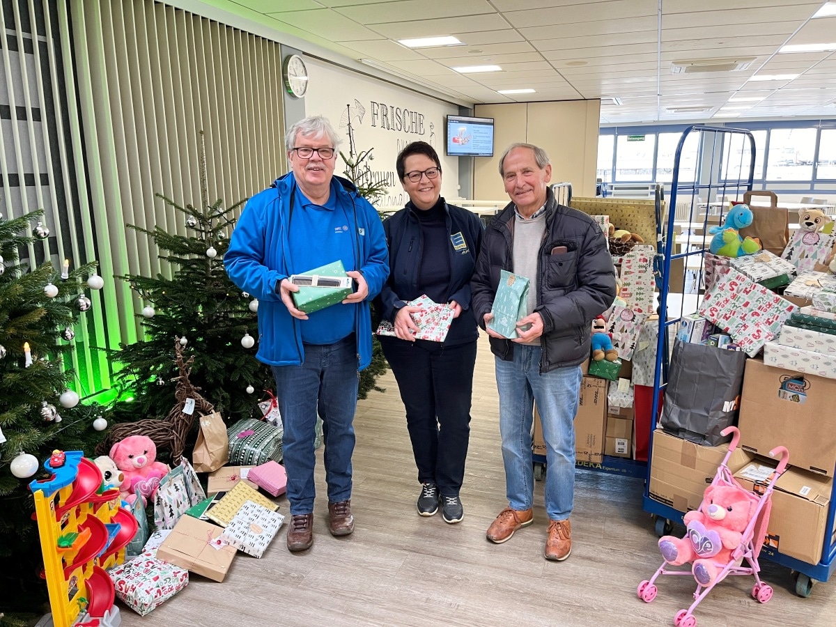 2 Männer und eine Frau mit Geschenken in der Hand, 2 Weihnachtsbäume und daneben 2 Rollbehälter voll mit Geschenken.