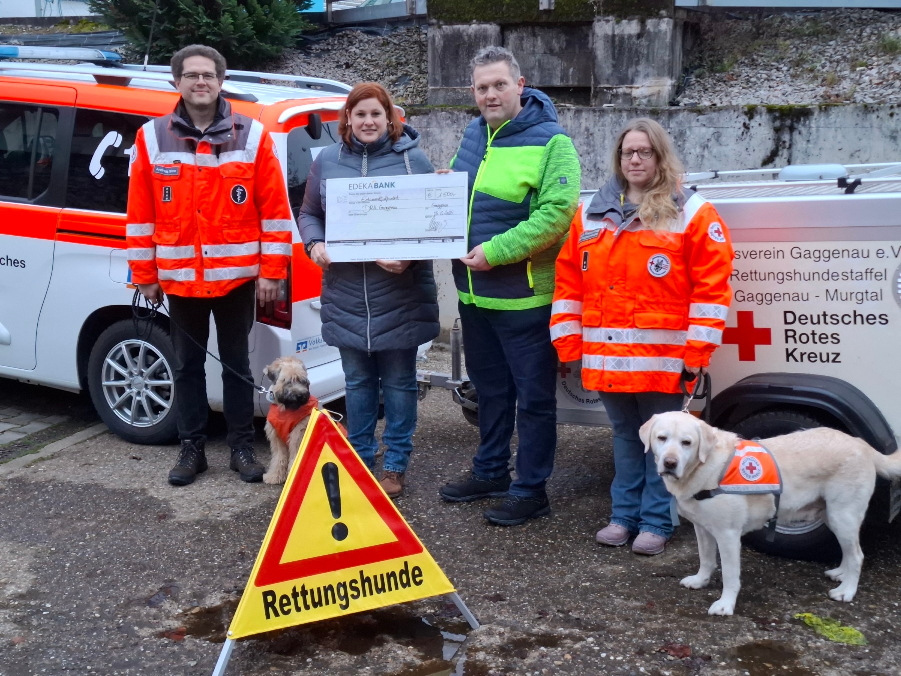 4 Personen (2 in Rettungsjacke) und 2 Rettungshunde posen mit einem Scheck und Warndreieck "Rettungshunde" vor dem DRK-Auto