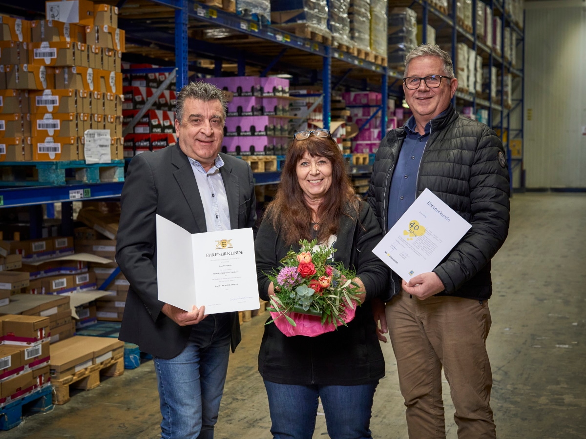 Eine Frau steht im Lager mit Blumenstrauß zwischen 2 Männern, die je eine Urkunde halten. Im Hintergrund sind Hochregale.