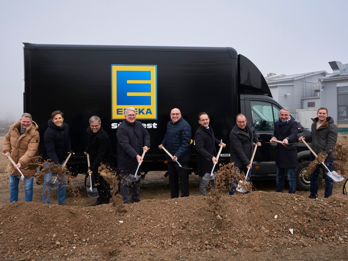 9 Männer mit Jacken und Schaufeln buddeln symbolisch beim Spatenstich vor einem schwarzen Transporter mit EDEKA-Logo.