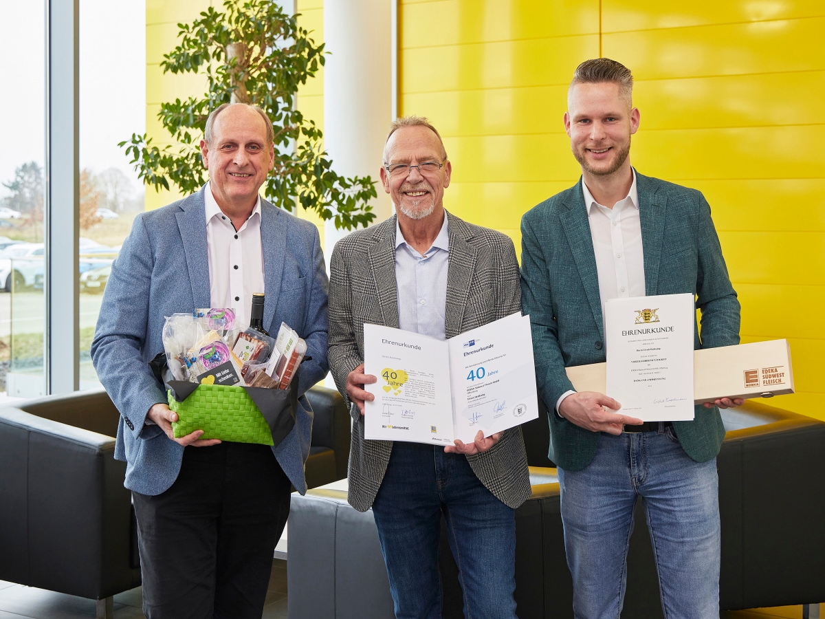 3 Männer stehen zusammen und halten einen Präsentkorb sowie 3 verschiedene Urkunden mit der Aufschrift 40 Jahre.