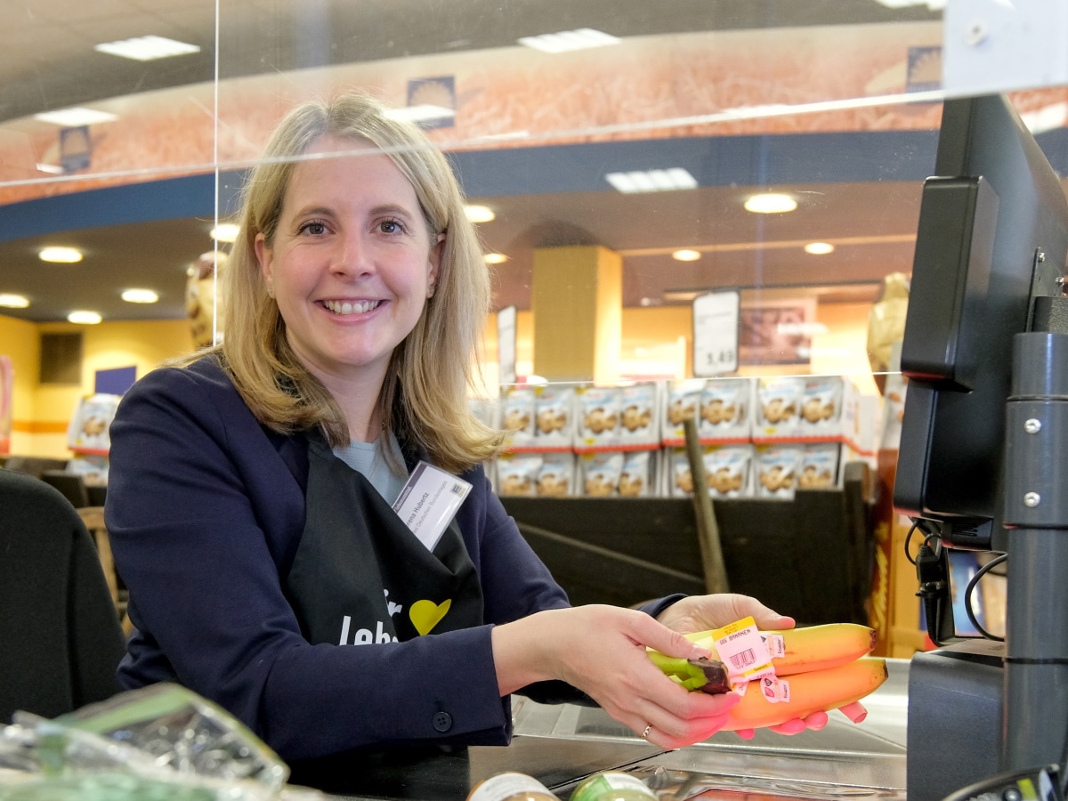 Eine blonde Frau sitzt an der Supermarktkasse, scannt Bananaen und lächelt in die Kamera.