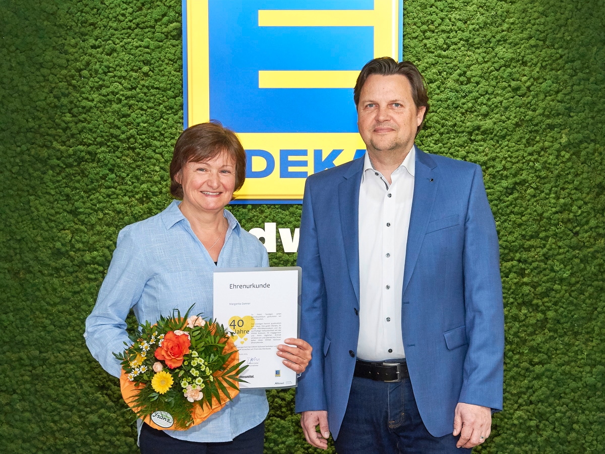 Eine lachende Dame steht mit Blumenstrauß und Urkunde nebem einem Mann vor einer moosbedeckten Wand mit EDEKA-Schild.