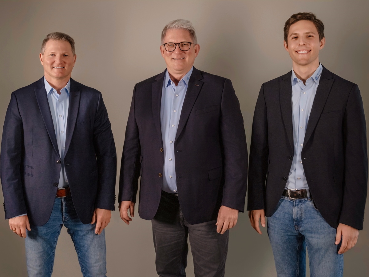 3 Männer in dunkelblauen Jackets und hellblauen Hemd stehen nebeneinander, mit Blick in die Kamera und lächeln. 