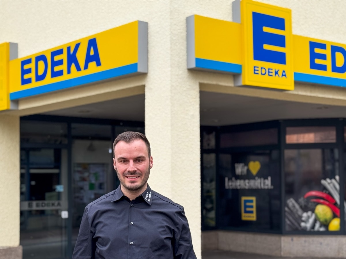 Ein Mann mit schwarzem Hemd steht vor dem Eingang eines Marktes an einer Hausecke mit EDEKA-Leuchtschildern darüber.