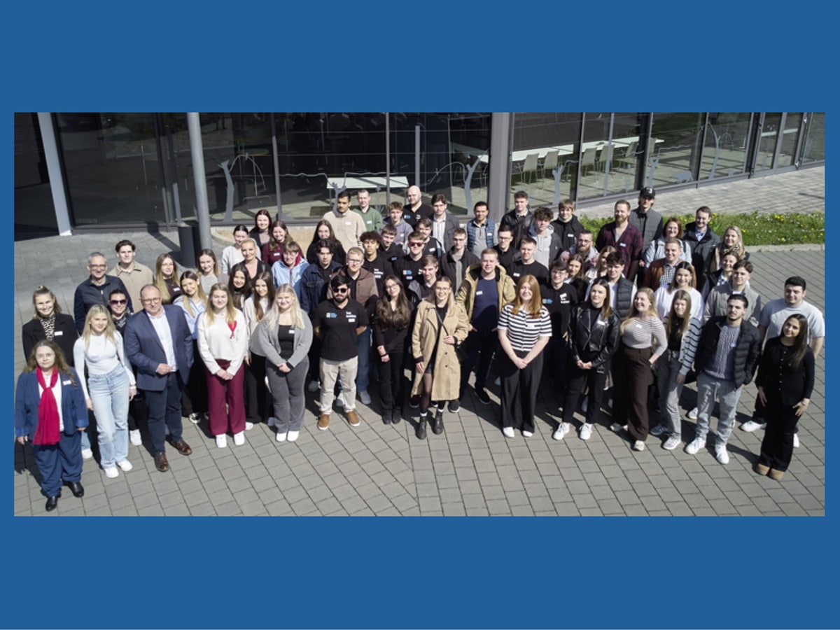 60 junge Frauen und Männer stehen für ein Gruppenbild bei Sonneschein zusammen und schauen in die Kamera.