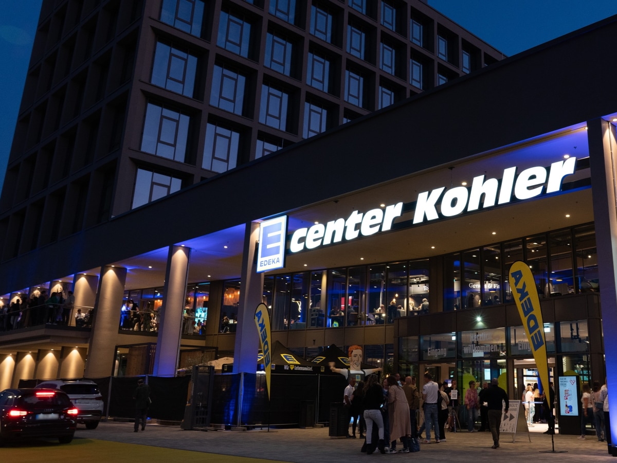 Bei Nacht leuchten Lichter und das Schild E center Kohler über dem Eingang. EDEKA-Flaggen und Menschen stehen draußen davor. 