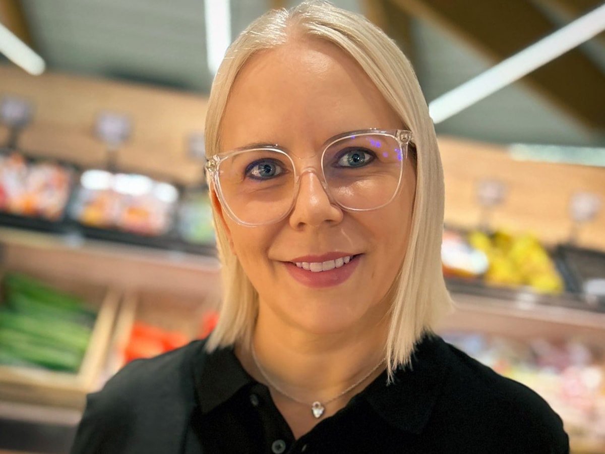 Ein Frau mit blonden schulterlangen Haaren und Brille lächelt in die Kamera. Im Hintergrund ist eine Gemüseabteilung zu sehn.