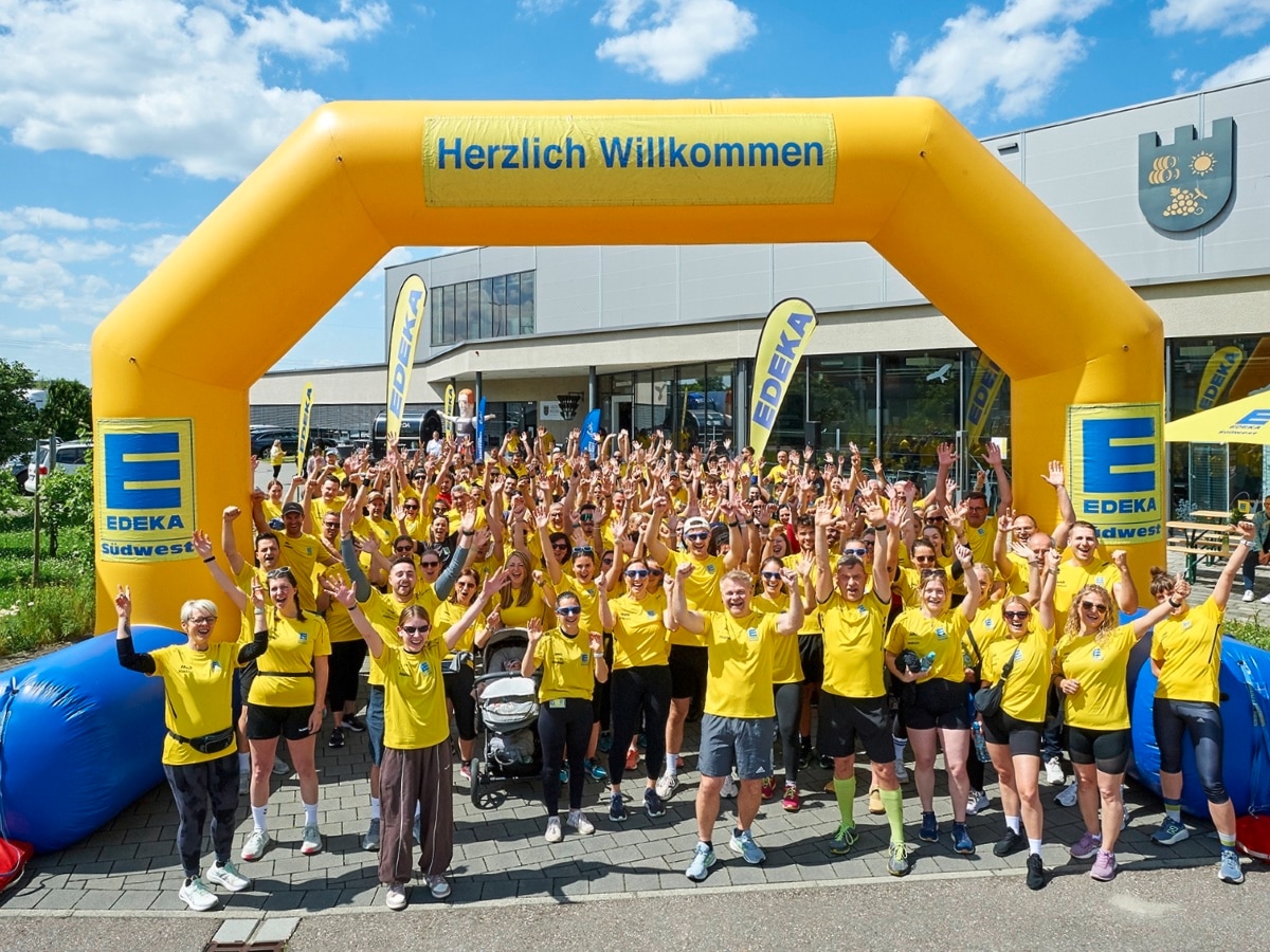 Rund 250 Läuferinnen und Läufer im gelben EDEKA T-Shirt jubeln unter einem gelben aufblasbaren Tor vor dem Start.