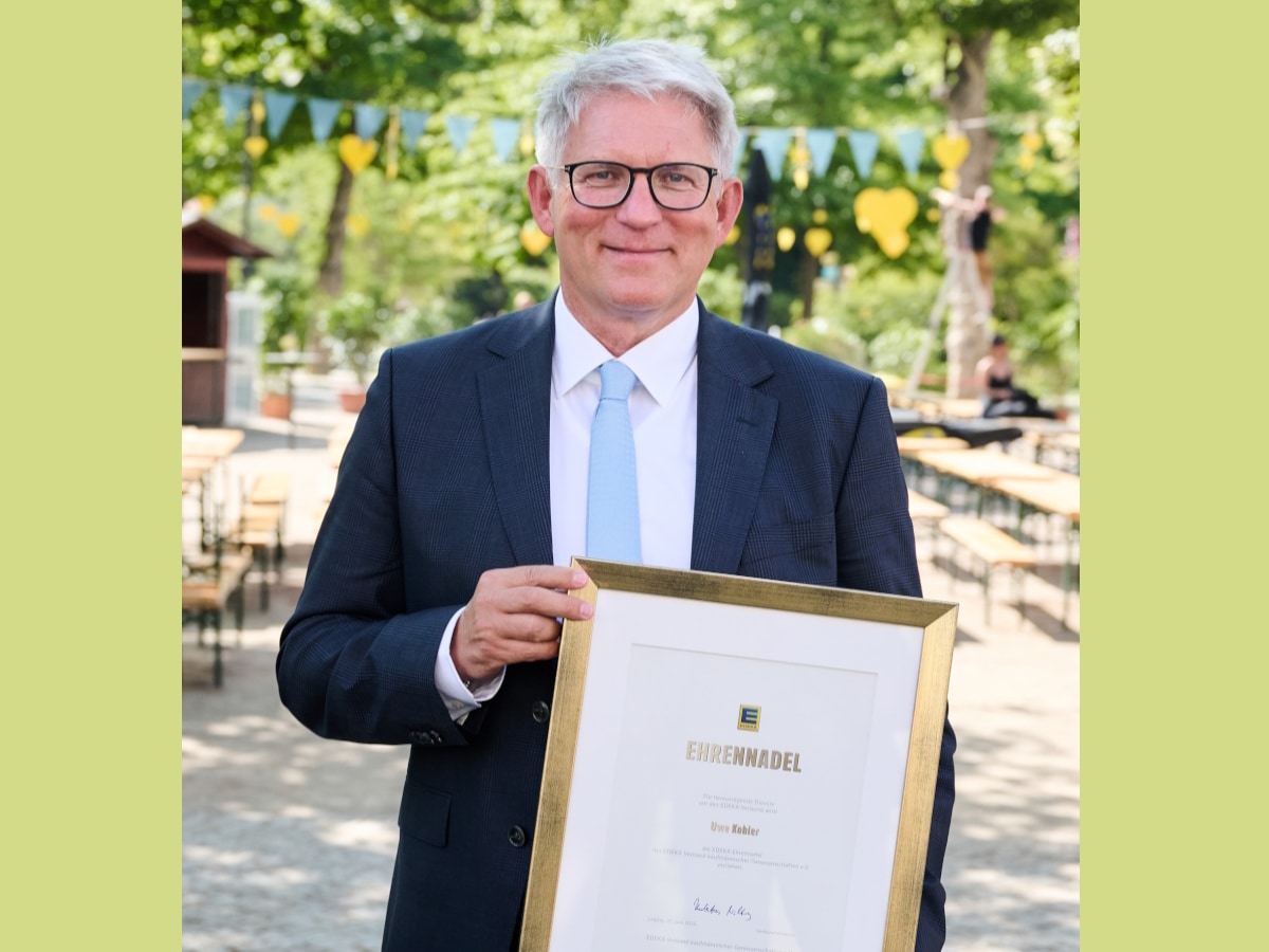 Lächelnder Mann mit grauen Haaren und blauem Anzug hält eine eingerahmte Urkunde mit der Aufschrift Ehrennadel in den Händen.