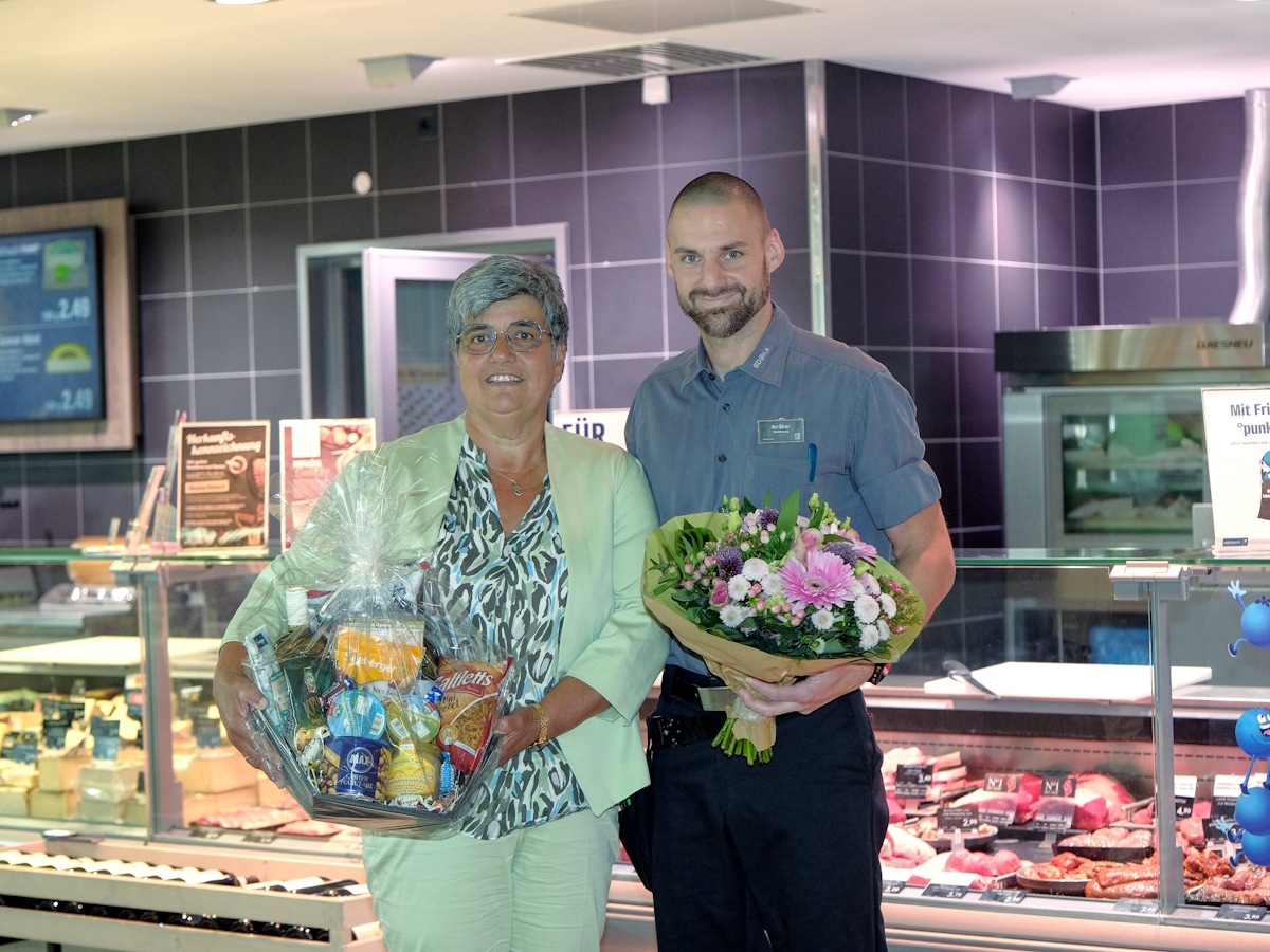 Vor der Bedientheke im Markt steht die Jubilarin mit einen Geschenkkorb, der Mann rechts neben ihr hält einen Blumenstrauß.