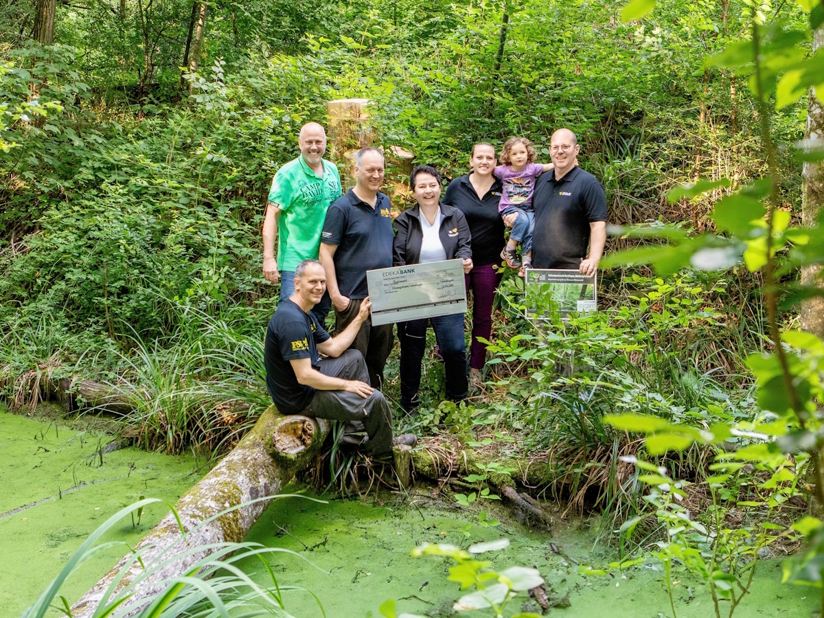 Eine Gruppe steht mit einem symbolischen Spendenscheck in einem saftig grünem Waldstück davor ein Gewässer und Wasserlinsen