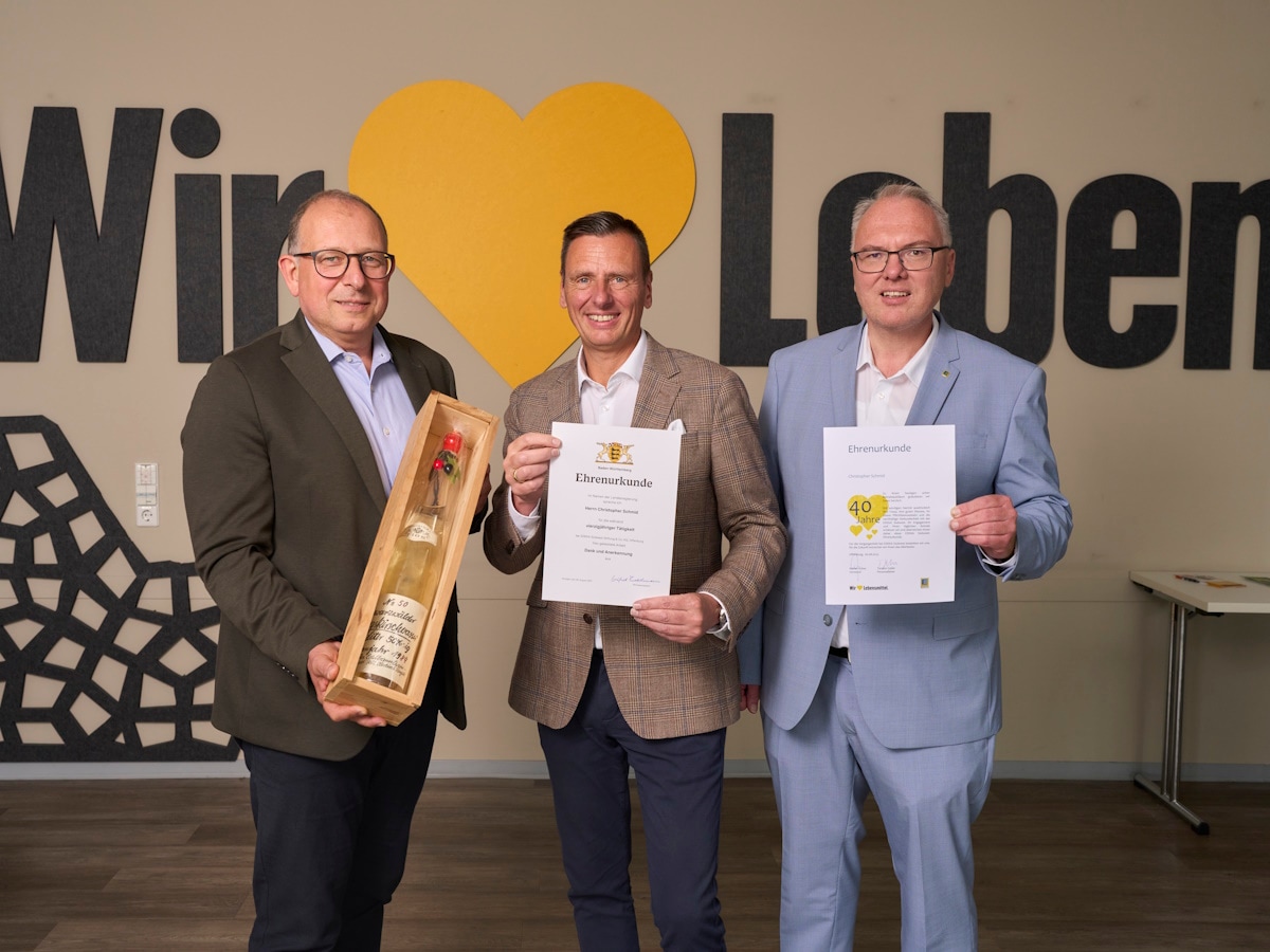 3 Männer stehen mit einem Weinpräsent und Urkunden vor einer Wand mit dem Schriftzug "Wir lieben Lebensmittel".