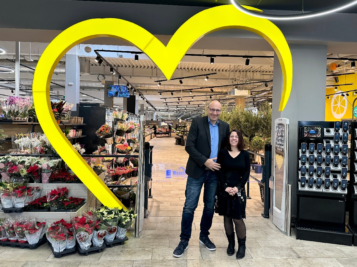 Ein Mann und eine Frau stehen vor im Eingang des Marktes vor einem großen gelben Herz.