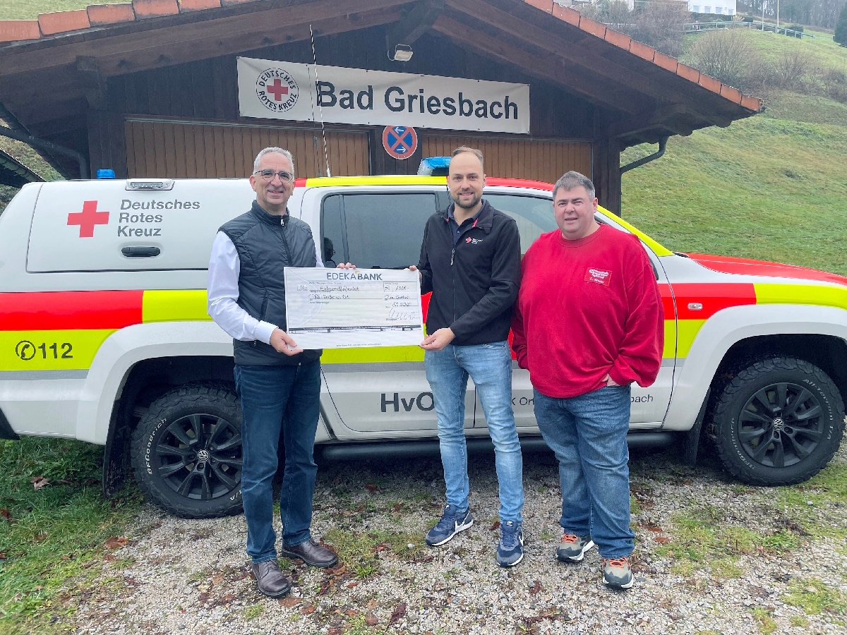 3 Männer stehen mit einem symbolischen Spendenscheck vor einer Hütte und einem DRK Auto.