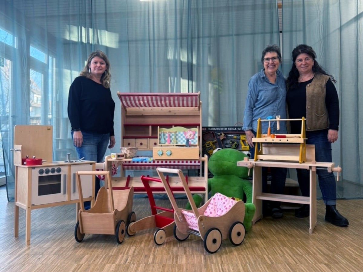 Neue Spielsachen aus Holz: Kinderküche, Kaufladen, Werkbank, Puppenwägen sowie ein großer Plüschfrosch, dahinter 3 Frauen
