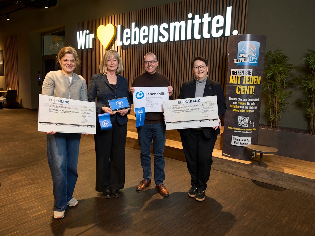 3 Frauen und 1 Mann stehen mit symbolischen Schecks vor einer Holzwand mit dem EDEKA-Slogan "Wir lieben Lebensmittel".