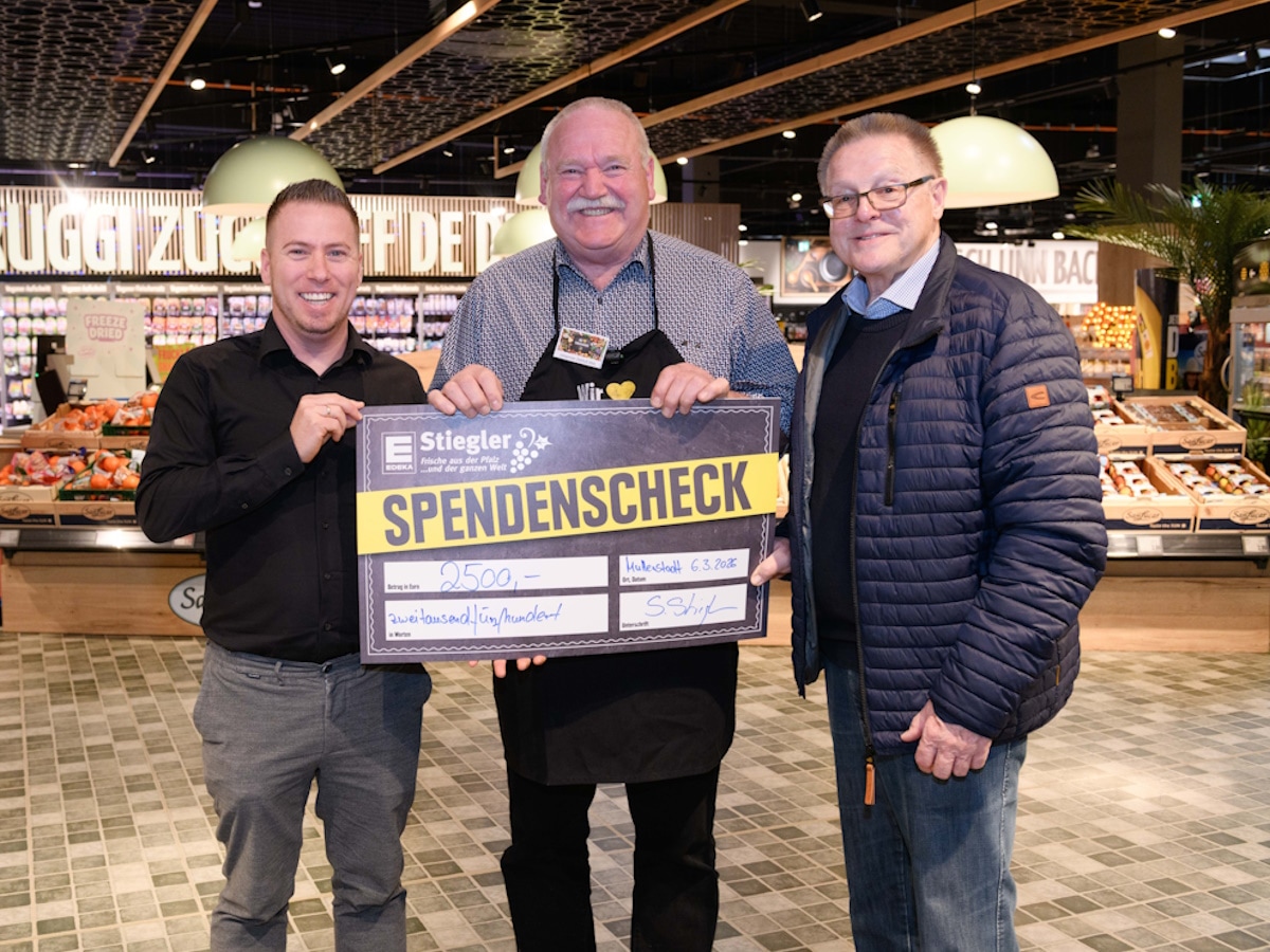 3 Männer stehen mit einem symbolischen Spendenscheck vor der Obst- und Gemüseabteilung.