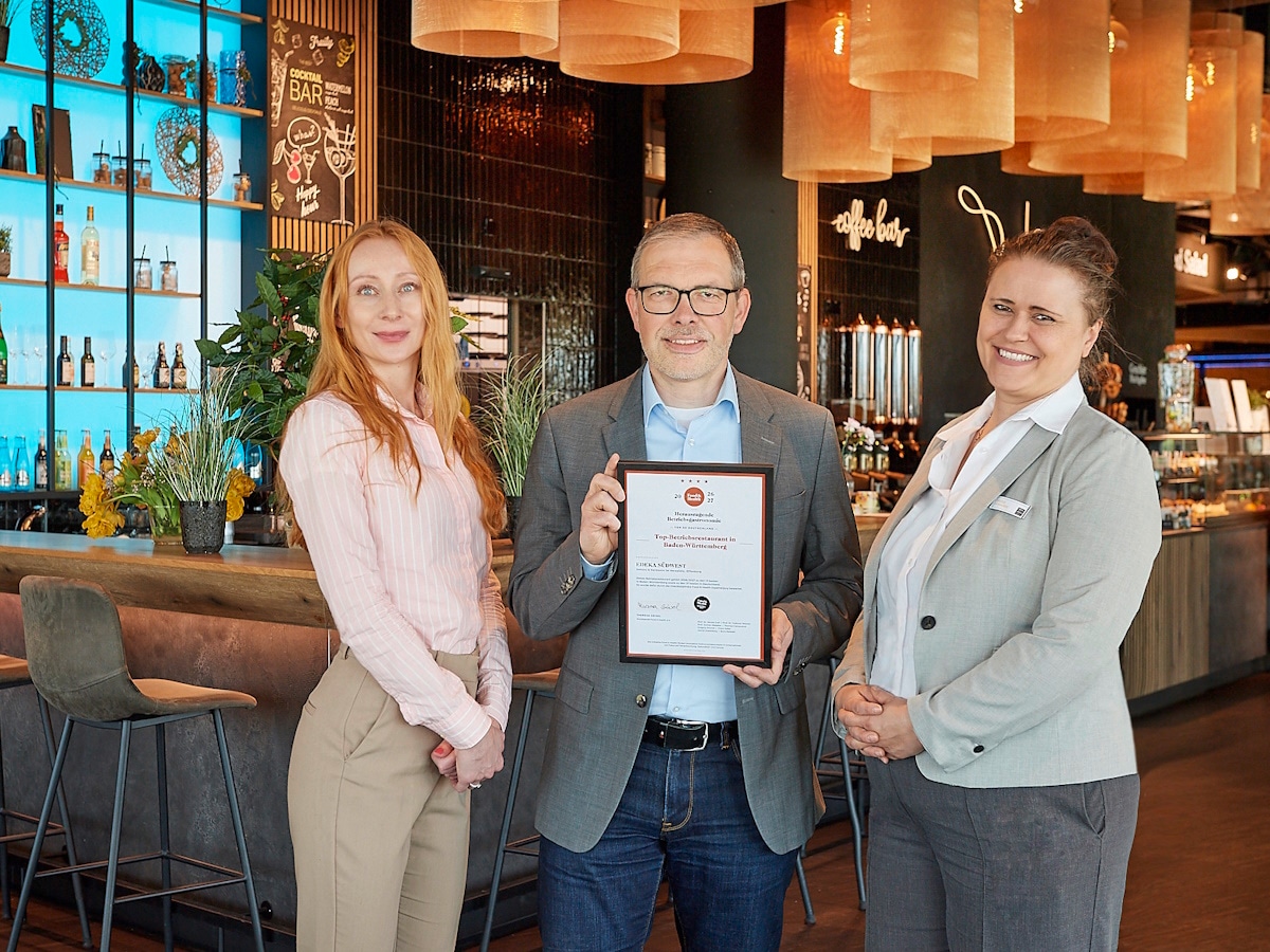 Ein Mann mit Urkunde steht in der Mitte zweier Frauen vor dem Betriebsrestaurant.