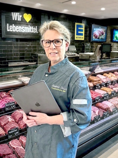 Ein Frau mit Tablet und Brille steht vor einer Fleischtheke im EDEKA-Markt