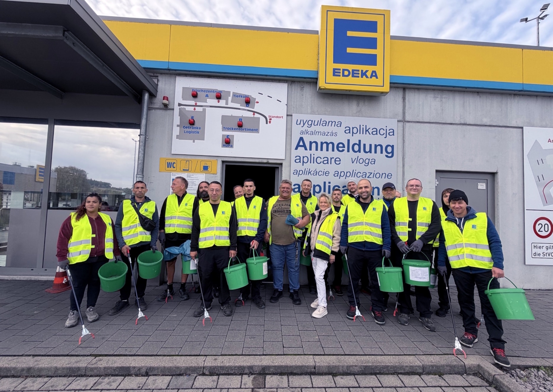 18 Mitarbeitende mit gelben Warnwesten, Eimern und Müllzangen stehen an der Pforte der EDEKA