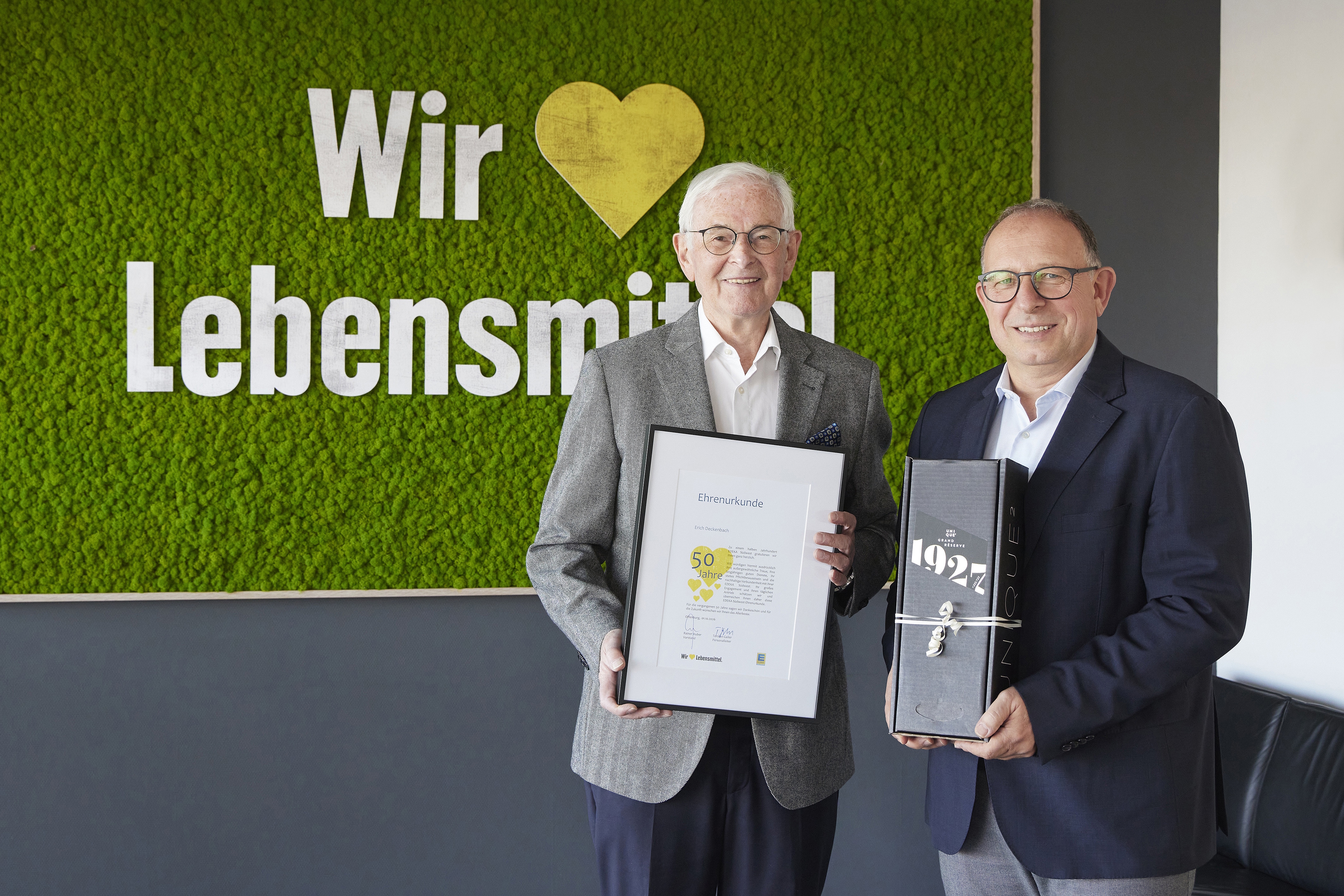 Der Jubilar hält eine Urkunde, daneben ein Mann mit einem Präsent. Dahinert der Claim "Wir lieben Lebensmittel."