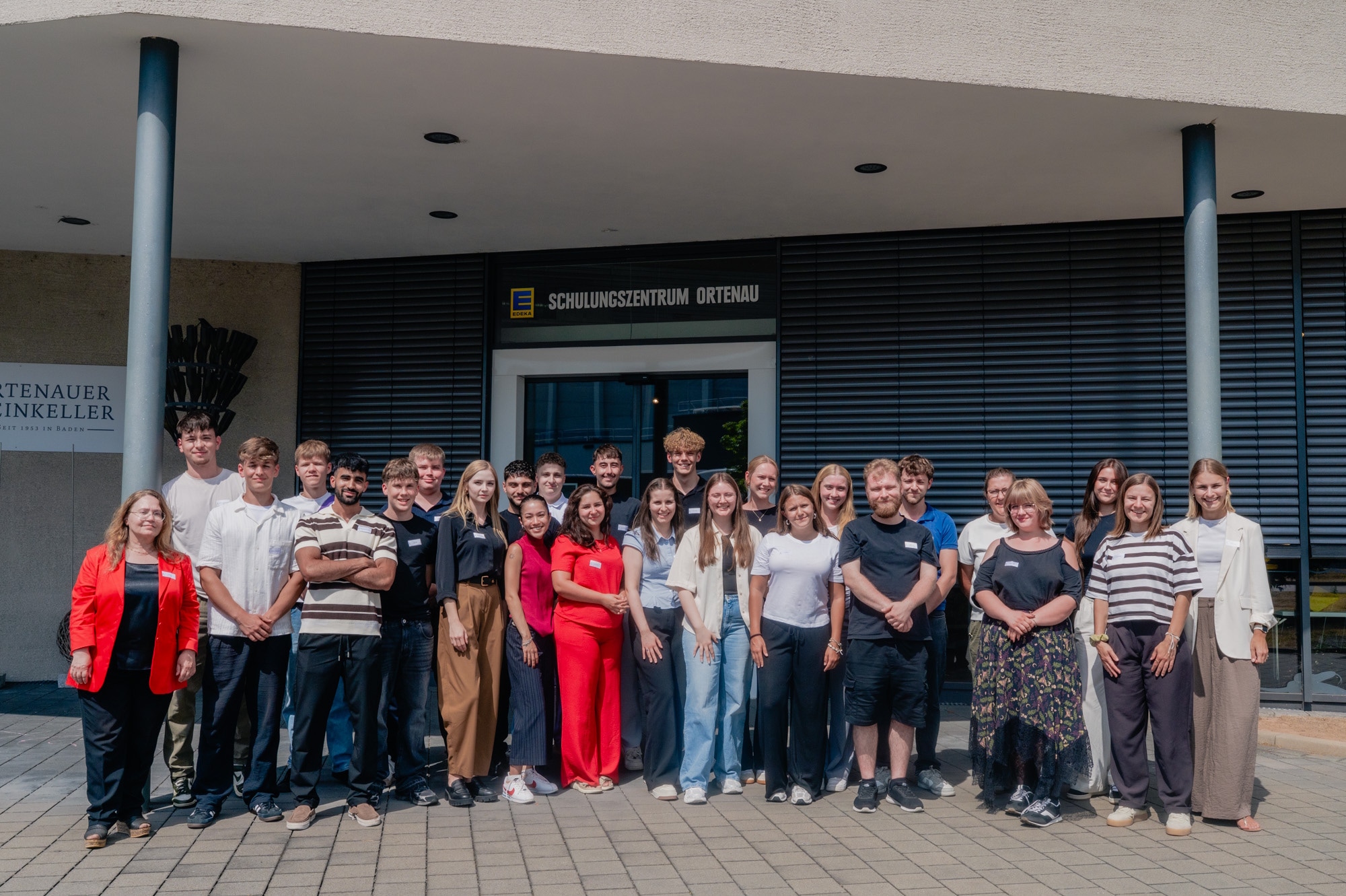 Eine Gruppe junger Menschen steht für eine Gruppenbild zusammen, sie schauen bei sonnigem Wetter in die Kamera und lächeln