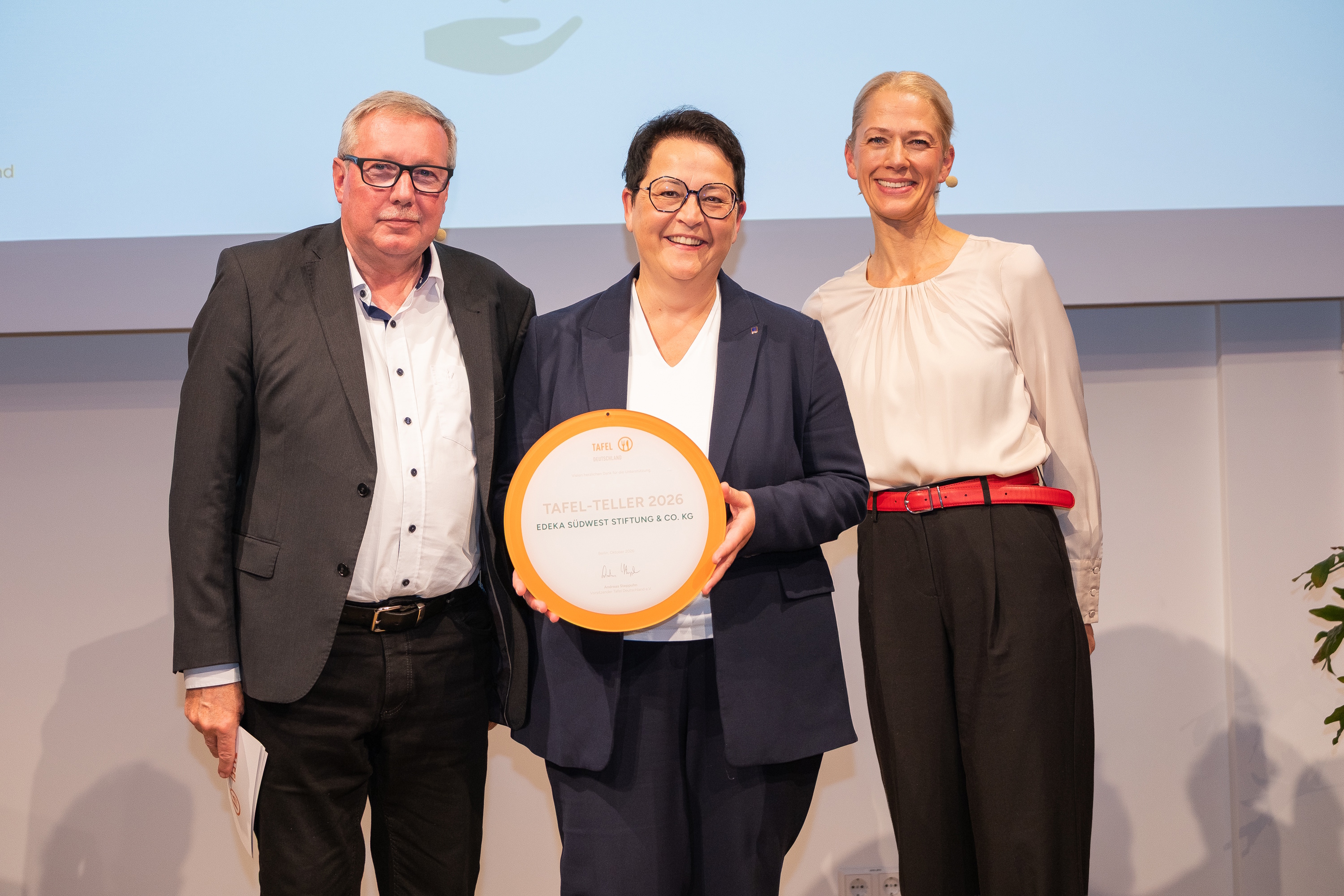 Ein Mann und 2 Frauen stehen nebeneinander auf der Bühne, die Dame in der Mitte präsentiert lächelnd einen Teller mit der Aufschrift Tafel-Teller 2026