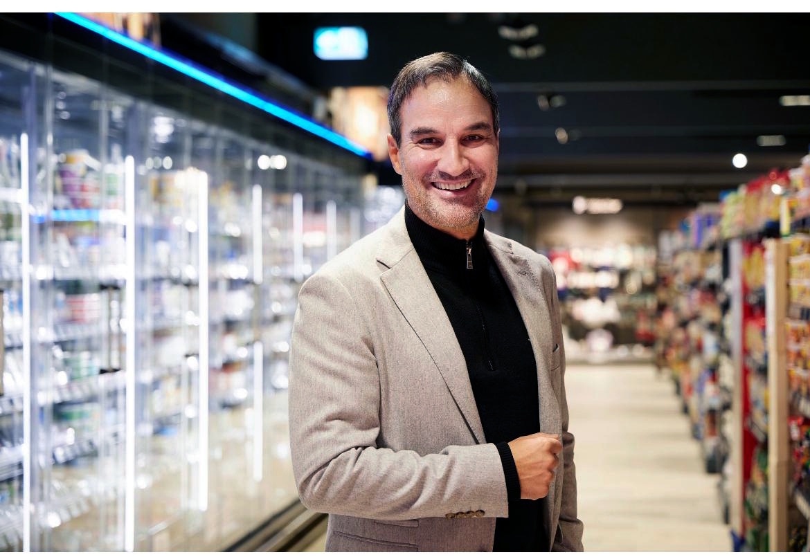 Ein Mann im Jackett steht zwischen den Regalen seines EDEKA Marktes und lächelt