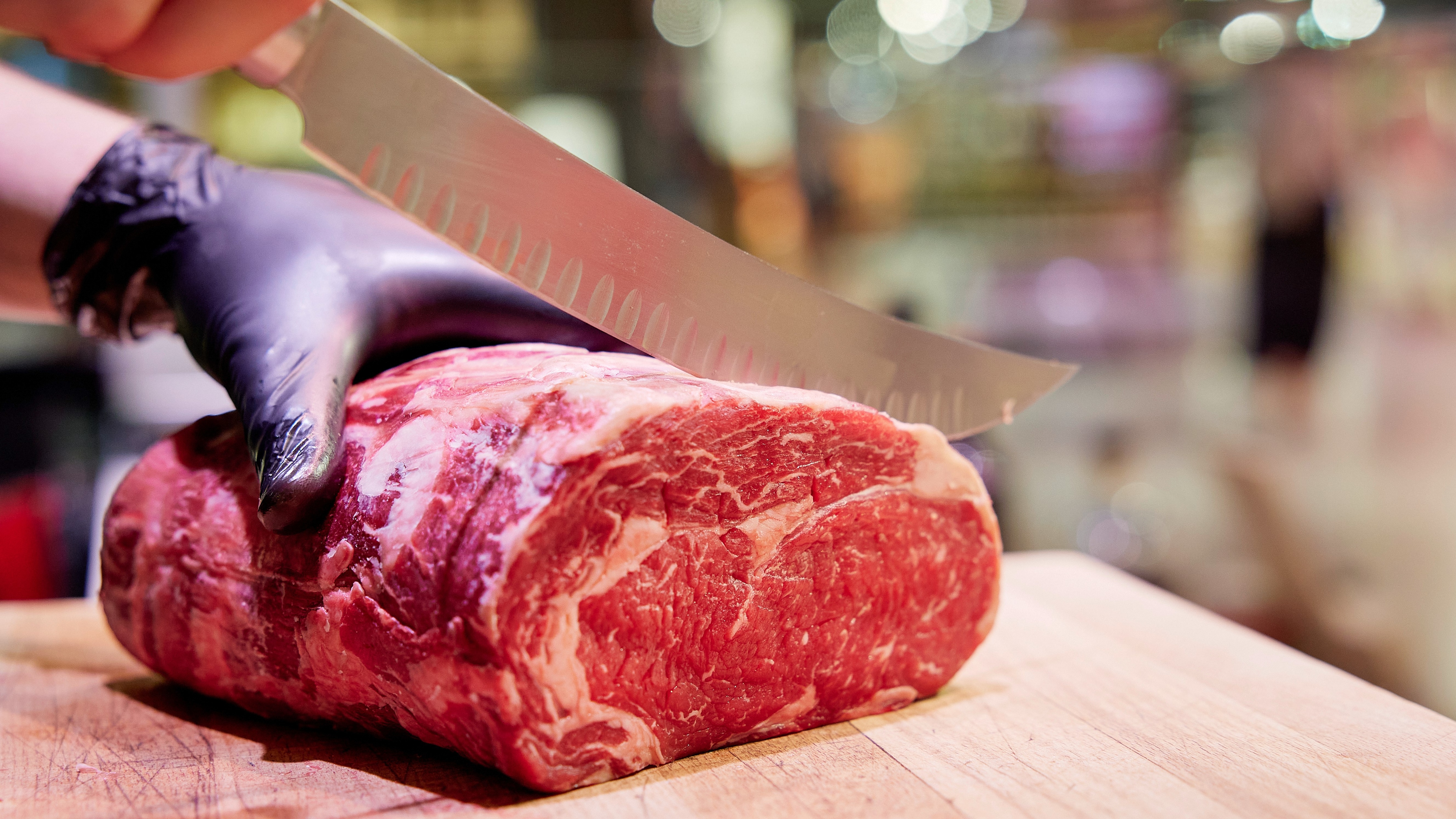 Ein großes Stück rohes Fleisch wird von einer Hand im Plastichandschuh gehalten. Ein Schlachtermesser schneidet von oben.