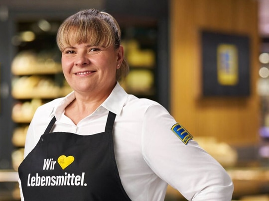 Lächelnde blonde Frau in weißem EDEKA-Hemd und schwarzer Schürze mit der Aufschrift Wir lieben Lebensmittel.