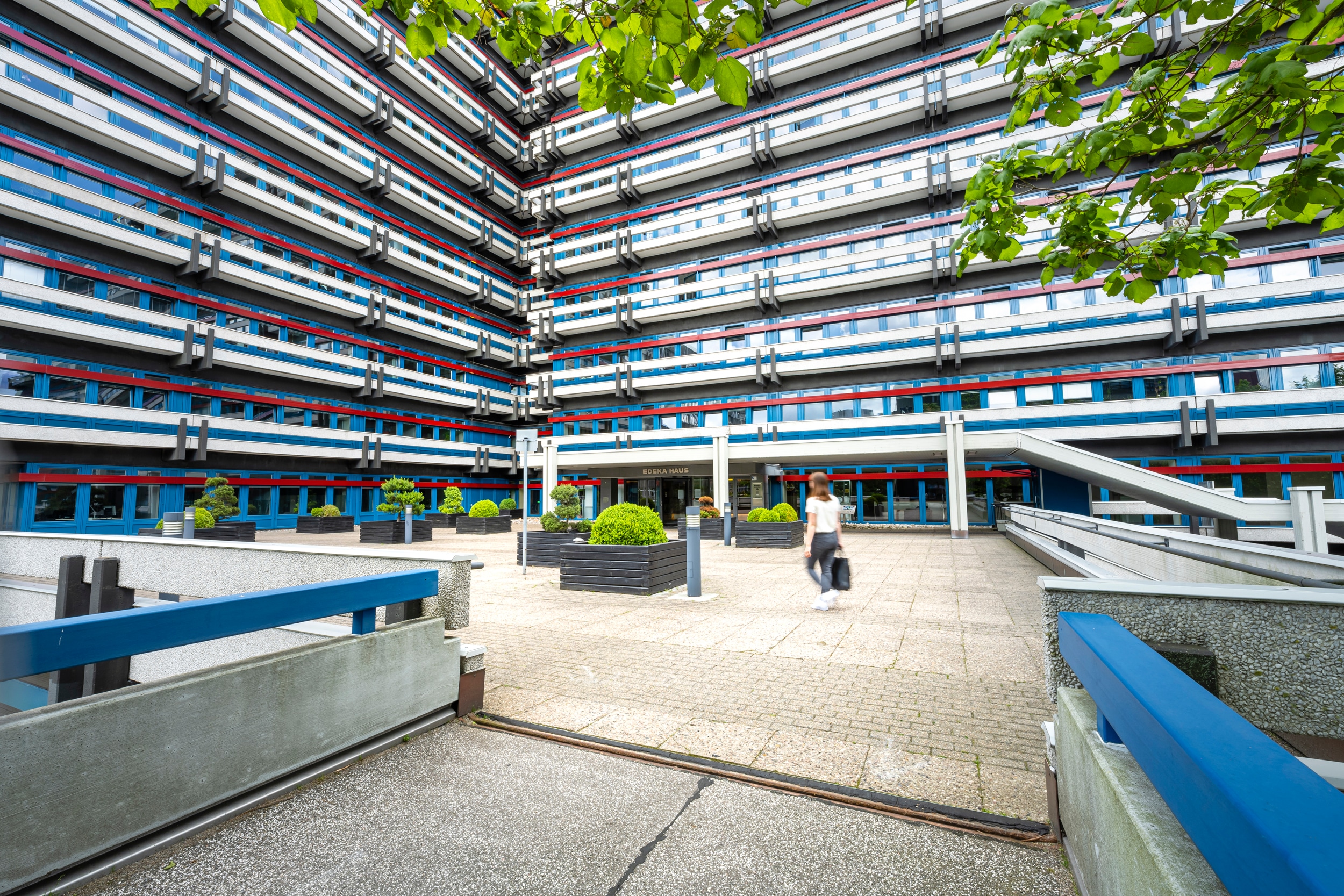 Ein Frau geht auf das Gebäude der EDEKA-Zentrale zu. Die Fassade ist blau mit roten und weißen Streifen.