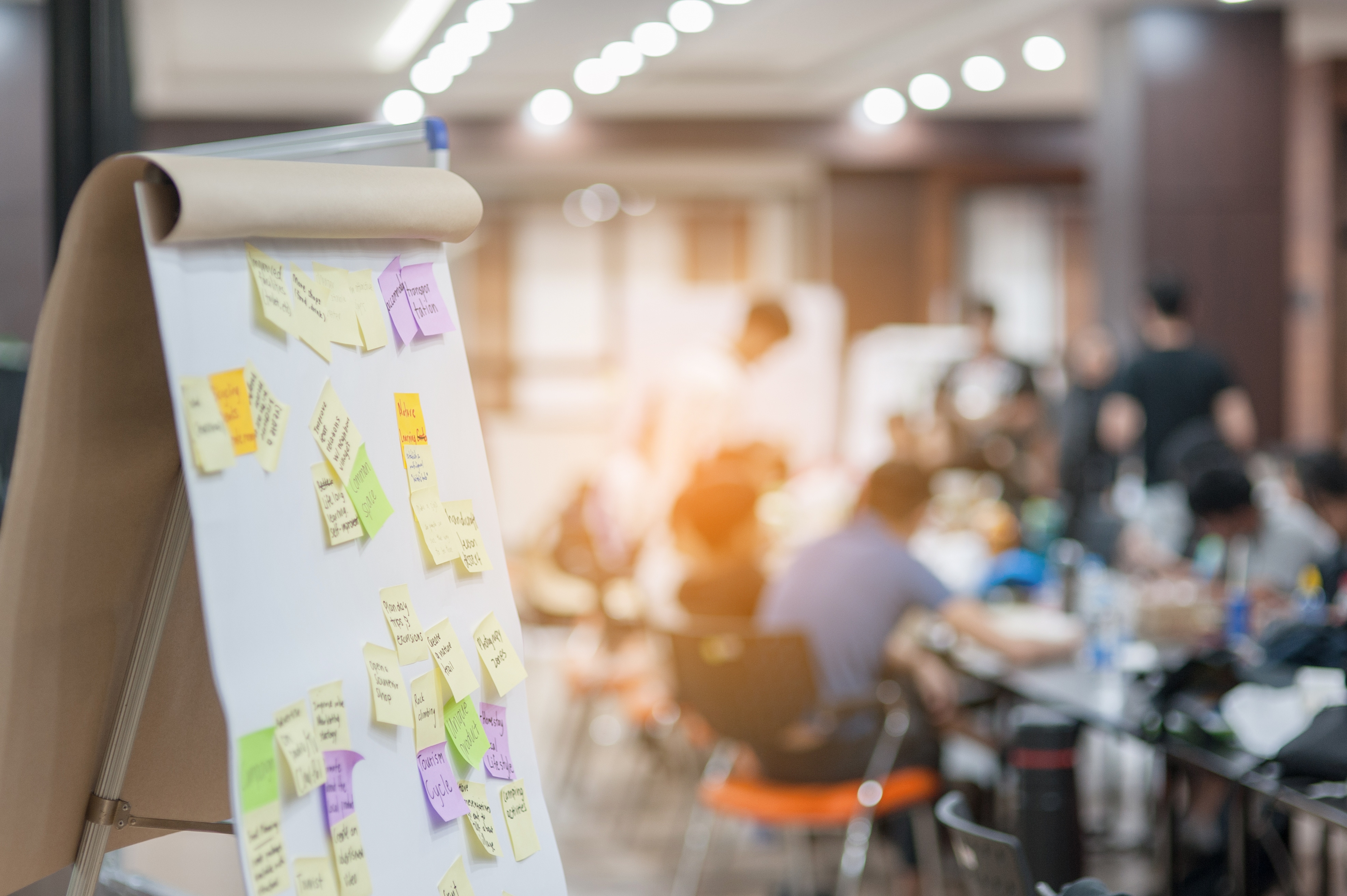 Flipchart mit bunten Post-its, im Hintergrund ist ein Meeting zu sehen. 