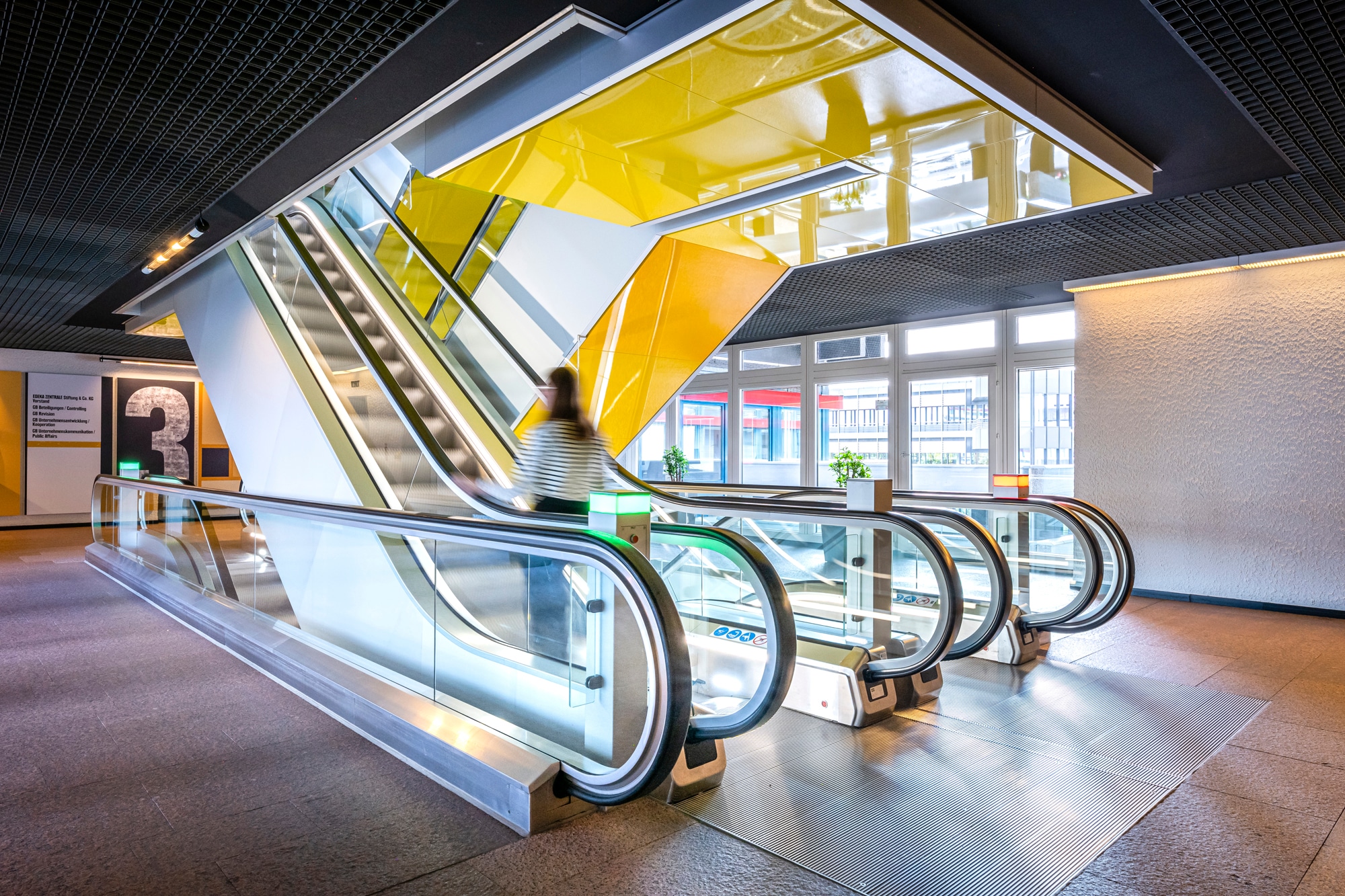 Rolltreppen in einem modernen Gebäude. Über den Rolltreppen ist gelbes Glas zu sehen. Eine Frau ist auf dem Weg nach oben. 