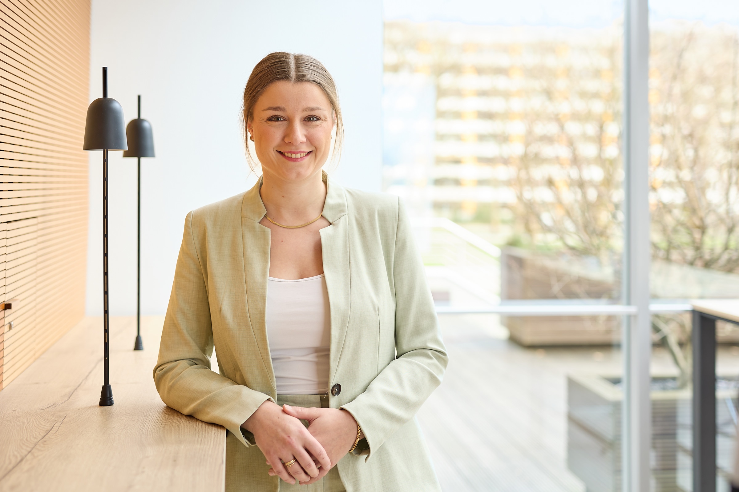 Düveke Düvel im Portrait, schaut lächelnd in die Kamera und lehnt am Holztisch