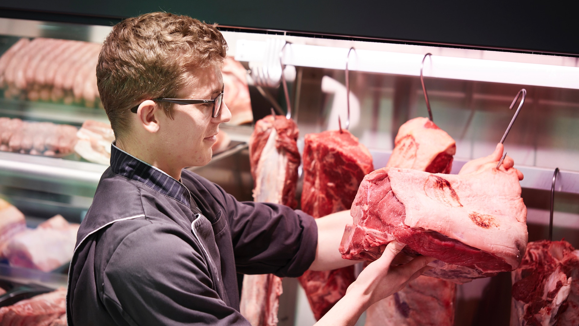 Ein Mann mit Brille und Schürze hält ein großes Stück Fleisch, das an einem Haken hängt.